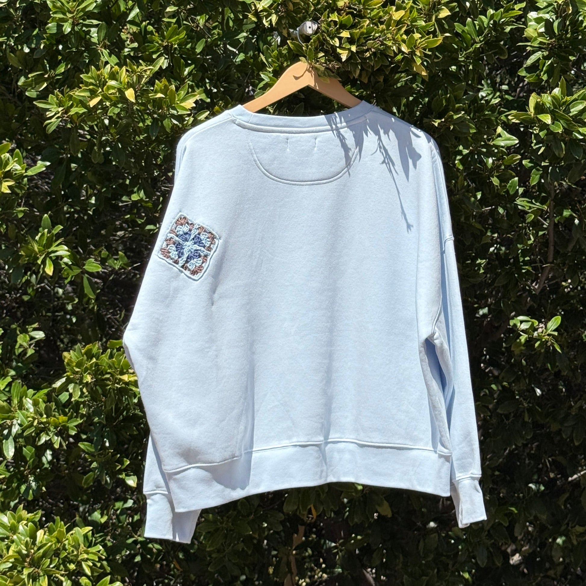 Light blue sweatshirt with floral designs on a hanger against a green leafy background