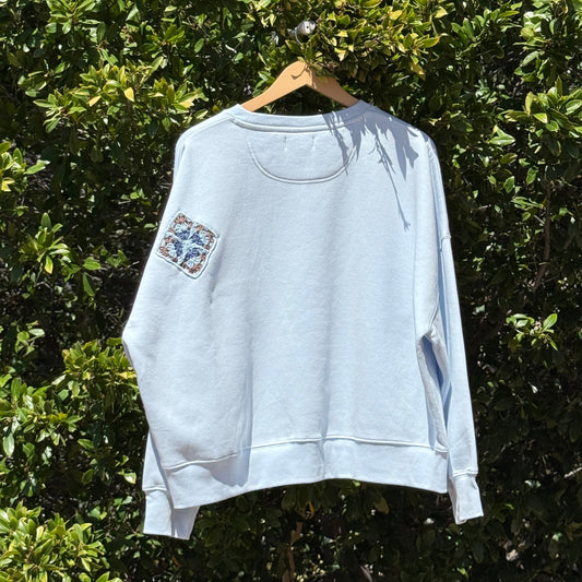 Light blue sweatshirt with floral designs on a hanger against a green leafy background