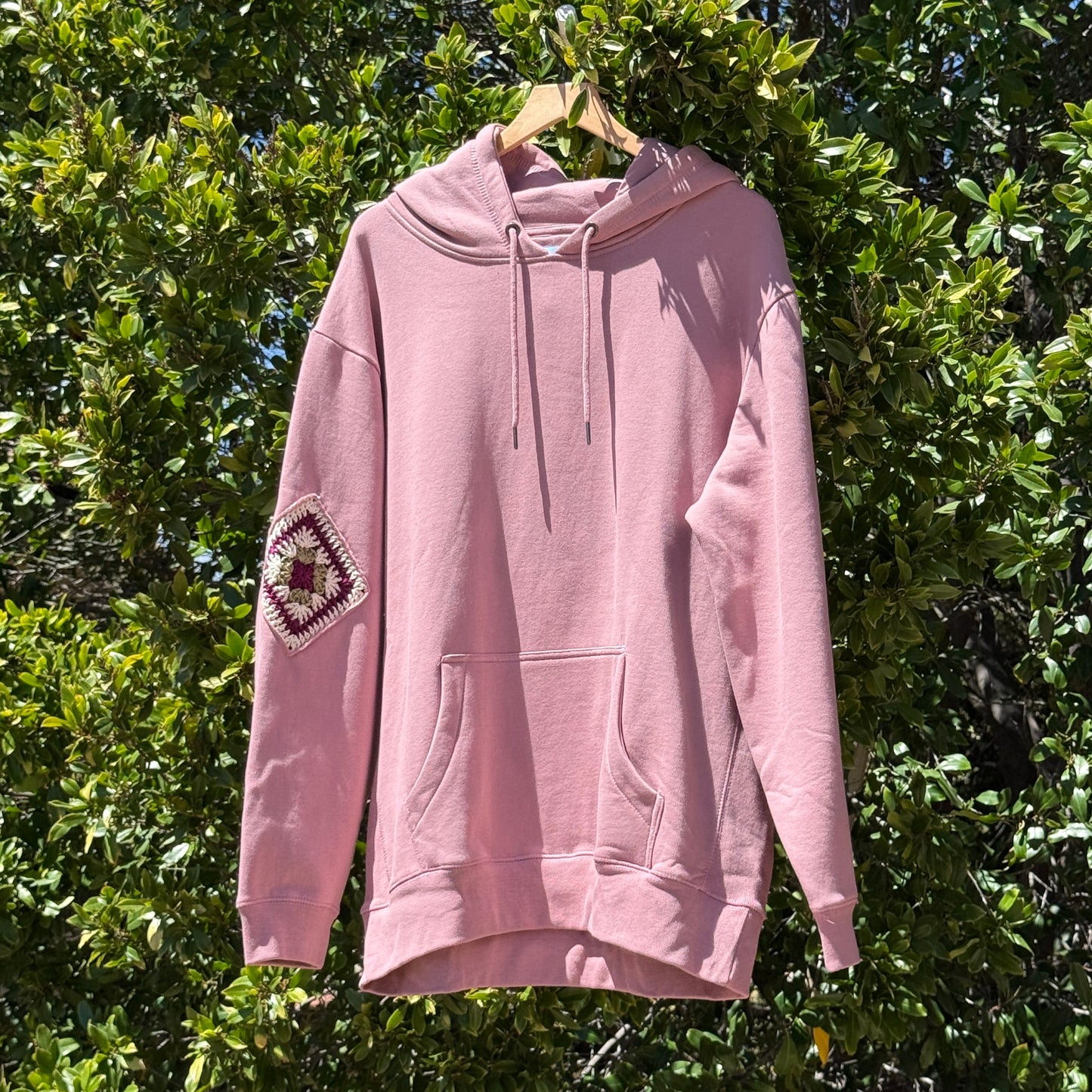 Pink hoodie with a colorful patch on the sleeve hanging against a green leafy background