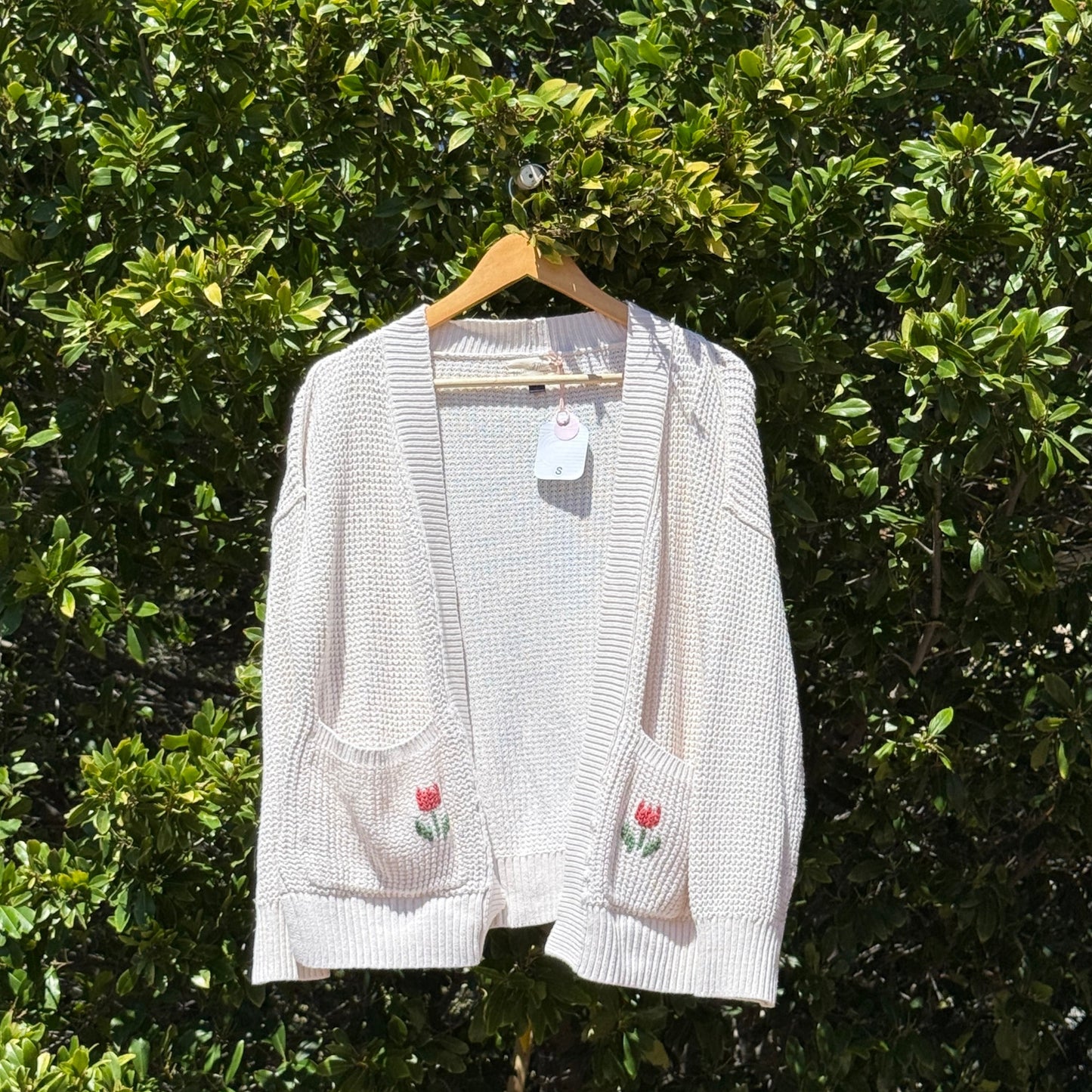 White cardigan with floral embroidery on a hanger against a green leafy background