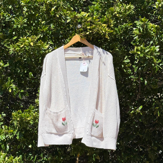 White cardigan with floral embroidery on a hanger against a green leafy background